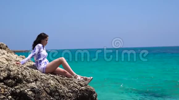 漂亮的女人坐在靠近水的石头上美丽的女人坐在海边的岩石上视频的预览图