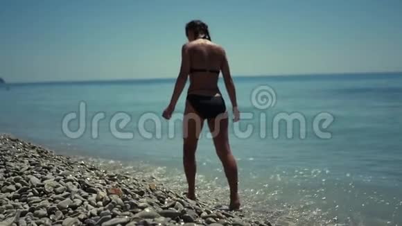 一个穿着泳衣的苗条女孩带着鹅卵石在蓝色的大海附近沿着海滩散步高清1920x1080慢动作视频的预览图