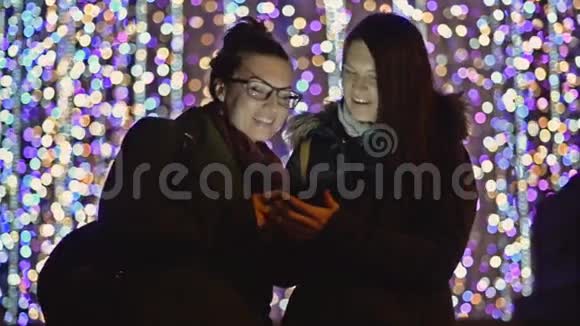 两个黑发少女笑着看圣诞灯光背景下的智能手机屏幕微笑女孩使用视频的预览图