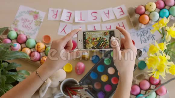 女人拿着智能手机用手工制作的装饰鸡蛋和字母制作桌子的照片复活节快乐视频的预览图
