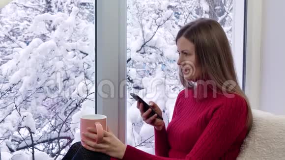 穿着红裙子的漂亮女孩坐在窗台上喝茶用智能手机冬天在外面视频的预览图