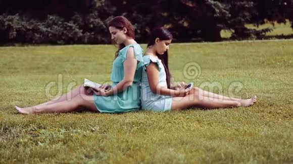 一个美丽的女孩和可爱的小女孩背靠背坐在草地上看书用平板电脑视频的预览图