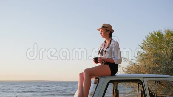 为年轻女性在靠近海堤的老式汽车上喝茶女孩手里拿着一杯咖啡视频的预览图