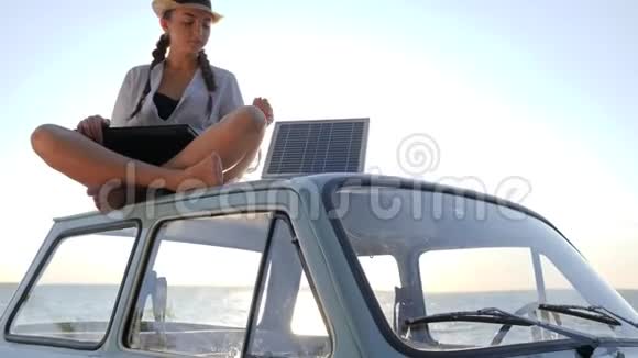 背光的露营女孩坐在车顶上带着太阳能阵列充电笔记本电脑挥手问好女孩子坐在老式车上视频的预览图