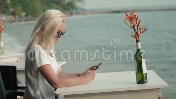 一个金发女孩在夕阳下的海边用手机在现代咖啡馆里微笑着的女人画像视频的预览图