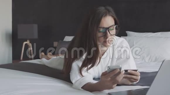 穿着白色浴袍和眼镜的女人用信用卡和智能手机在线购物躺在酒店的床上视频的预览图