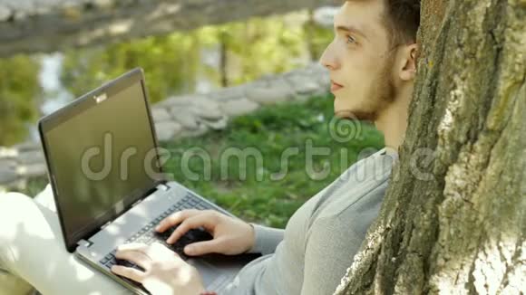 英俊的男子坐在树下的阴影与笔记本电脑和思考视频的预览图