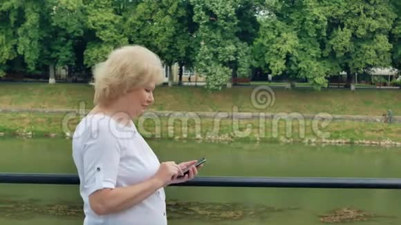 老年妇女沿着海滨散步使用带有耳机的智能手机河流的背景视频的预览图