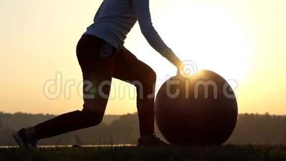苗条的女人站着跪着在夕阳下滚动她的健身球视频的预览图