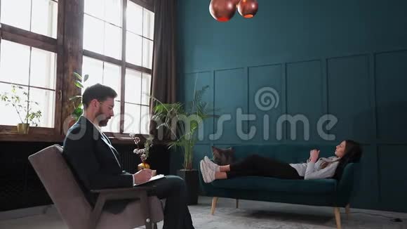 年轻女性在豪华的蓝色心理治疗中在舒适的沙发上看男心理学家视频的预览图
