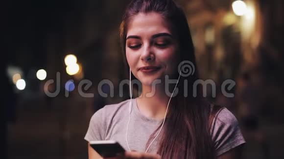 迷人的年轻女性年轻女性耳朵里有耳机听音乐使用智能手机模糊的夜晚视频的预览图