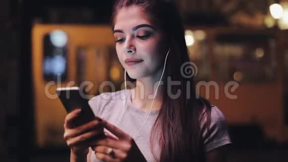 迷人的年轻女性年轻女性耳朵里有耳机听音乐使用智能手机模糊的夜晚视频的预览图