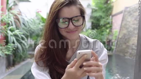 戴眼镜的女人微笑着在热带国家的游泳池附近使用智能手机视频的预览图