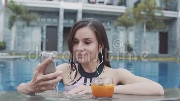 年轻的女人在泳池里用可尾音乐听着来自smarthone的耳机视频的预览图