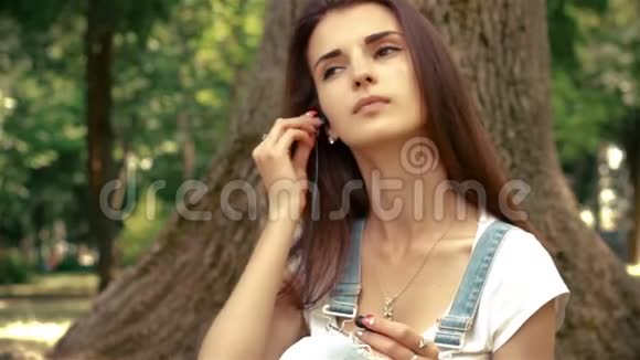 美丽的小姐耳朵里戴着耳机坐在树旁听音乐视频的预览图