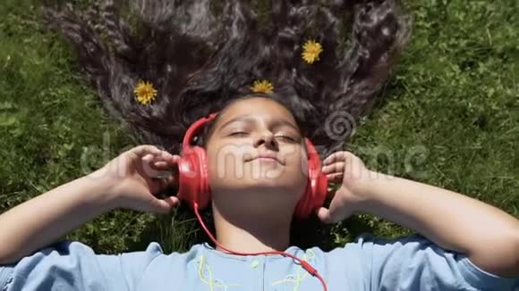 迷人的女孩留着长长的黑发躺在公园的草地上听着耳机上的音乐慢动作视频的预览图