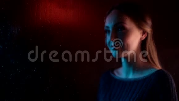 年轻悲伤的梦中女子靠近蒸汽玻璃窗在蓝色的霓虹灯中触动心灵身后是一个女孩视频的预览图