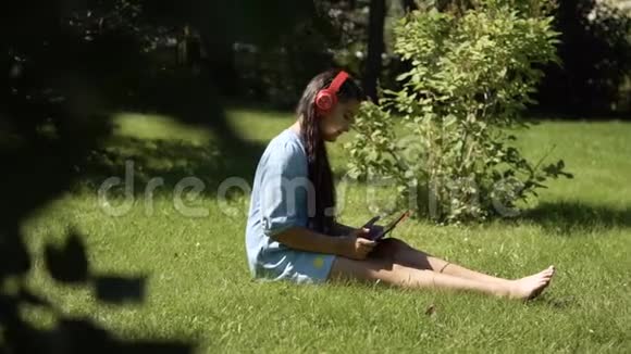 坐在公园草地上的平板电脑用耳机听音乐的女孩视频的预览图