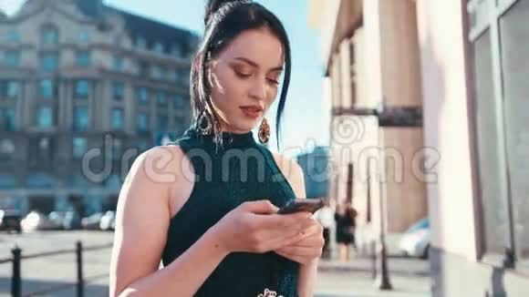 美丽的年轻女子在明亮的阳光下使用她的手机而站在市中心高兴地发短信抚摸视频的预览图