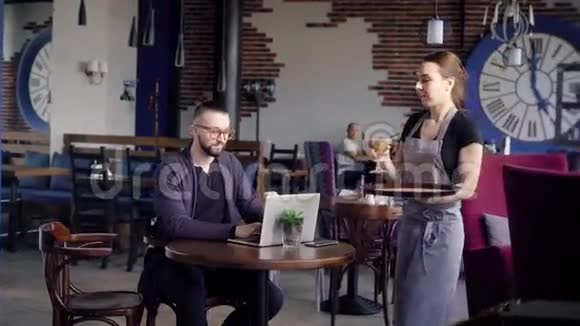 女服务员给商人端来一杯咖啡或卡布奇诺在桌子上笔记本电脑和手机一个年轻人视频的预览图