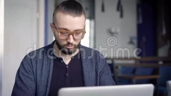 在桌子旁的手提电脑上工作的商人一个戴眼镜的年轻人看起来像个时髦的人随便看看视频的预览图