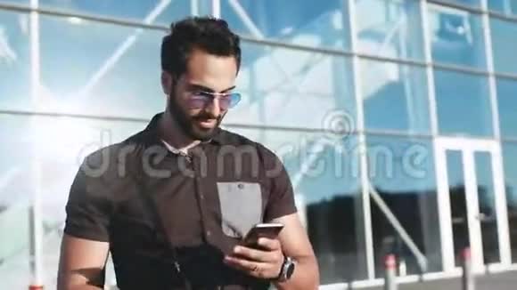 一个戴着太阳镜的年轻大胡子壮汉离开机场用他的手机高兴地对它微笑社会网络视频的预览图