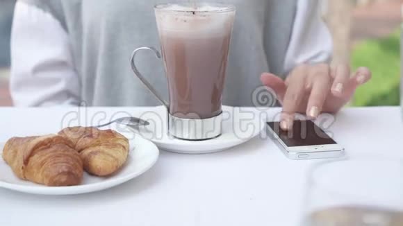 年轻女子一边喝着牛角面包一边看着智能手机上的照片女性使用手机经商视频的预览图