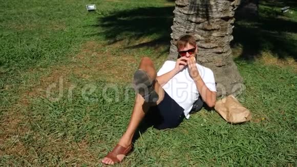 很有魅力的人坐在公园树下的草地上吃一个新鲜的汉堡4K慢动作视频的预览图