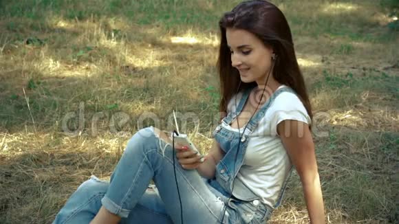 美丽的微笑女孩坐在街上的草地上听音乐视频的预览图