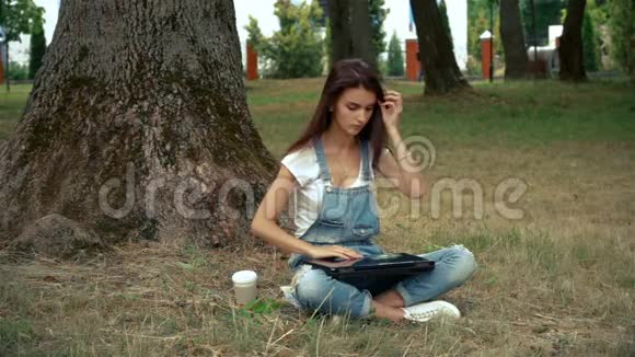 夏天坐在树旁的可爱女孩在笔记本电脑上打印视频的预览图