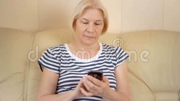 开朗好看的高级女性坐在家里的沙发上利用她的智能手机浏览视频的预览图