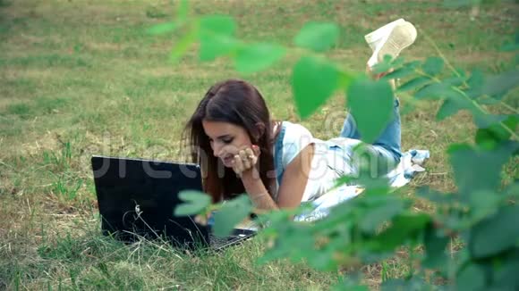 可爱的女孩穿着笔记本电脑躺在草地上的公园里视频的预览图