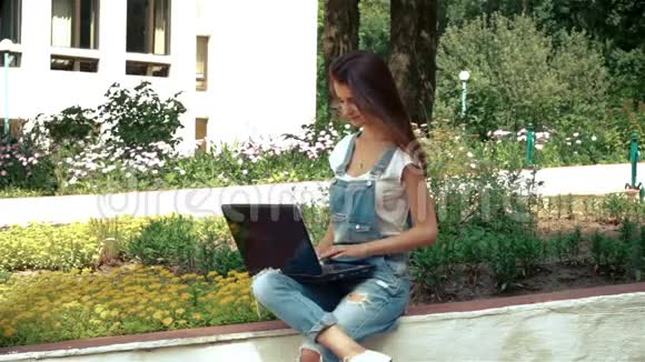 穿着牛仔裤连身裤的可爱的黑发女人坐在街上看着笔记本电脑视频的预览图