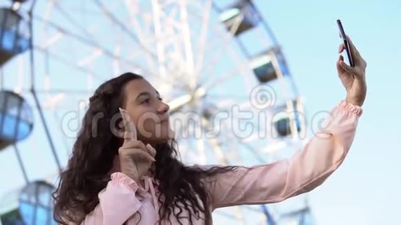 一个穿着连衣裙留着长发的女孩用手机在摩天轮附近自拍慢动作视频的预览图