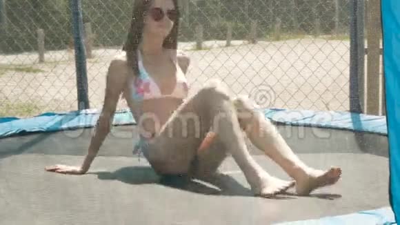 穿着泳衣和眼镜的有趣的黑发女人躺在蹦床上视频的预览图
