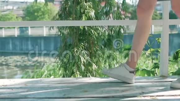 穿着白色运动鞋女孩的脚是在绿色的柳叶背景下的白色木板上视频的预览图
