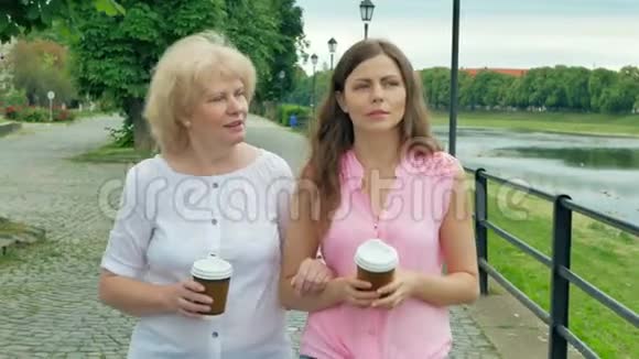 快乐的老妇人和年轻女子沿着海滨散步喝咖啡成年母亲和女儿在公园散步视频的预览图
