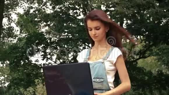 美丽的年轻女子坐在树旁手提电脑头发飞舞视频的预览图