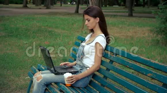 年轻女孩在公园里用你的腿拿着笔记本电脑喝咖啡视频的预览图