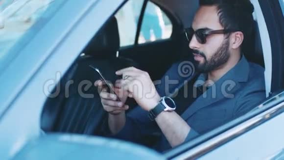 英俊的微笑男人戴着时尚的太阳镜坐在豪华的黑色汽车里用他的手机在等一个人视频的预览图