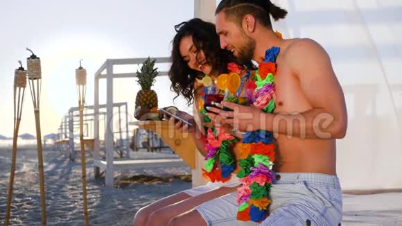 移动通信女孩坐在海滩上手里拿着手机漂亮的夫妇年轻人视频的预览图