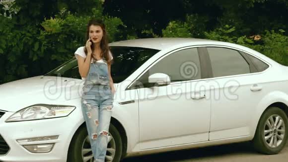 穿着牛仔裤的迷人年轻女性在她的车旁打电话视频的预览图