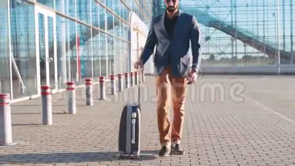 自信英俊的胡子男穿着时髦的西装把手提箱拉到机场航站楼商务旅行视频的预览图
