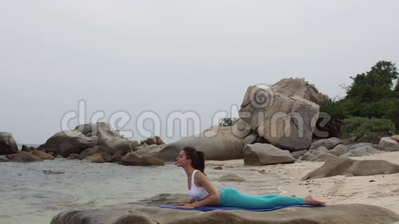 日落时分年轻女子在海滩上练习瑜伽体式眼镜蛇的姿势视频的预览图