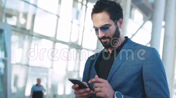 站在机场门口的英俊留胡子的东方商人用他的电话和微笑高兴地接待了视频的预览图