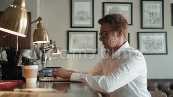 年轻的男性自由职业者在自然美食咖啡馆里坐在打开的笔记本电脑前一边用手机聊天视频的预览图