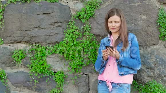 穿着棉夹克的漂亮女孩用智能手机带耳机背景中野生石头的复古墙视频的预览图