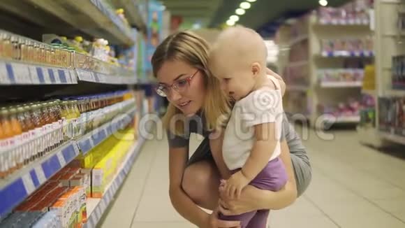 年轻漂亮的妈妈戴着眼镜和她可爱的孩子一起在超市的货架上挑选婴儿食品视频的预览图
