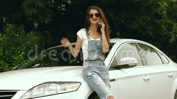 戴着镜子眼镜的女士站在白色汽车旁边用手机说话视频的预览图