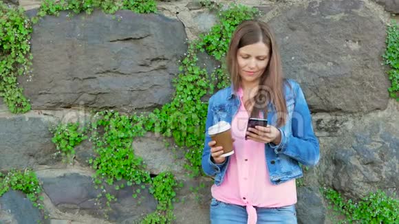 穿着棉袄用智能手机喝咖啡的漂亮女孩背景中野生石头的复古墙视频的预览图